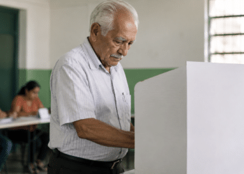 Homem idoso de cabelos brancos votando em urna branca em sala de votação com mesárias ao fundo