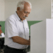 Homem idoso de cabelos brancos votando em urna branca em sala de votação com mesárias ao fundo
