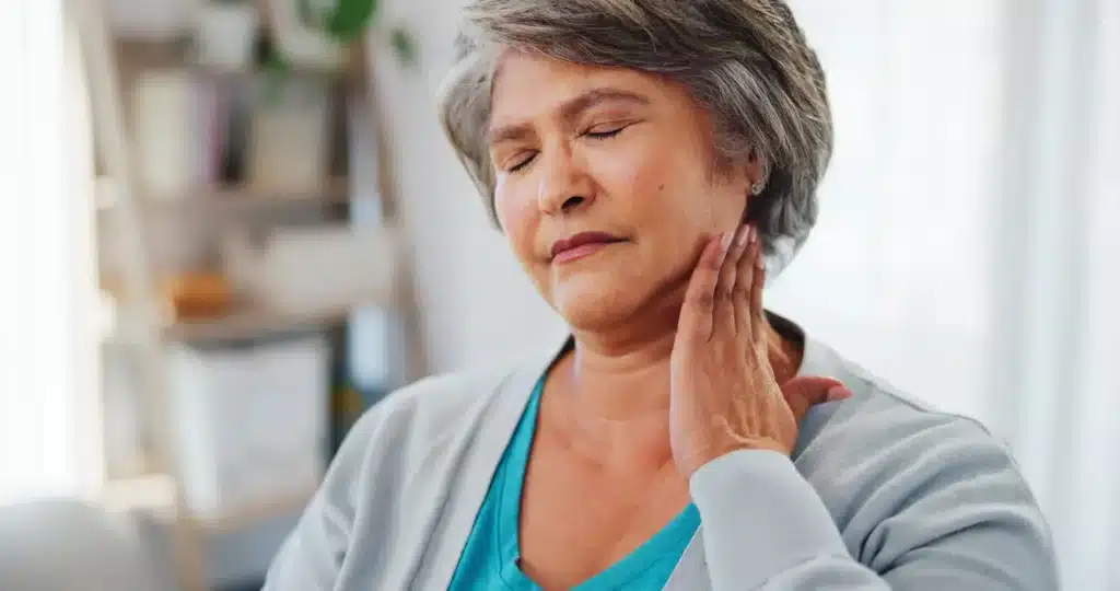 Mulher idosa toca o pescoço com expressão de desconforto, representando sinais de câncer de cabeça e pescoço.