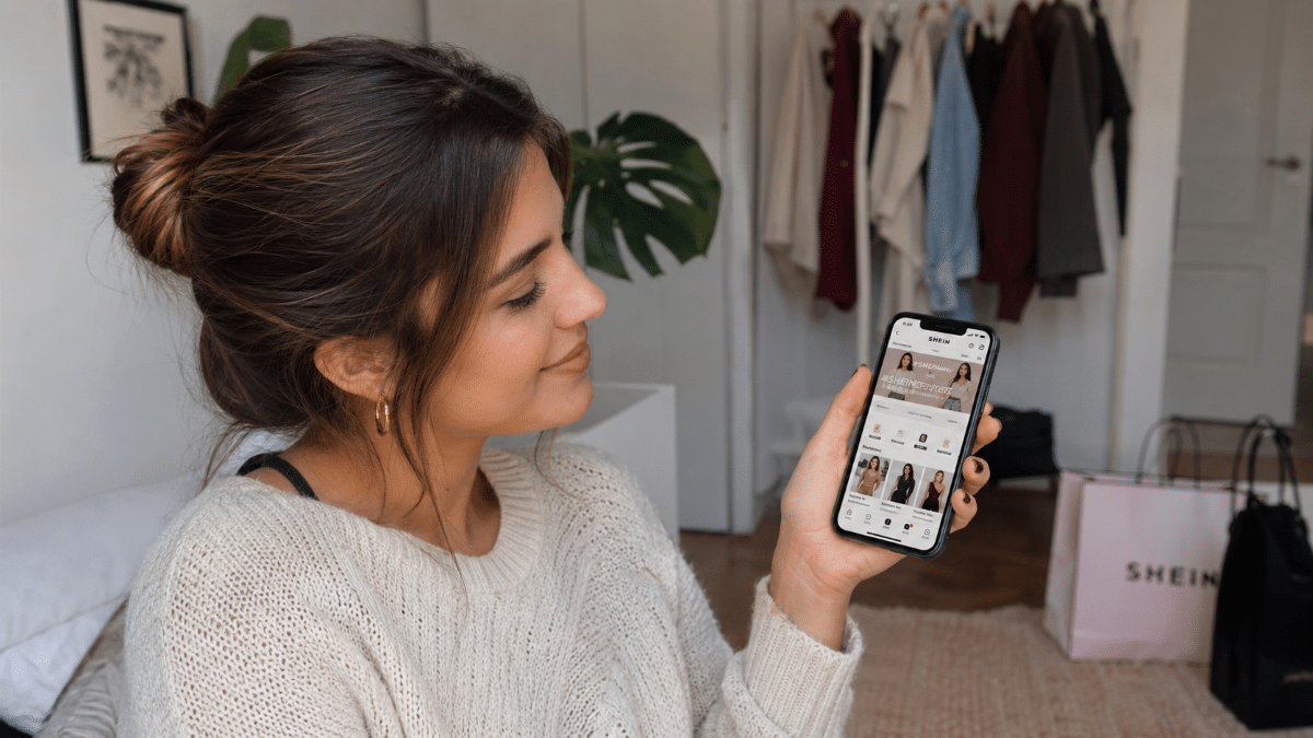 Mulher jovem com cabelo preso acessa o app SHEIN no celular em quarto com sacola da marca ao fundo