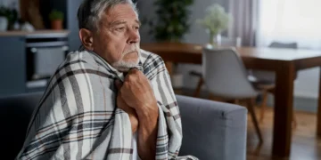 Idoso enrolado em cobertor sente frio enquanto está sentado no sofá de casa.