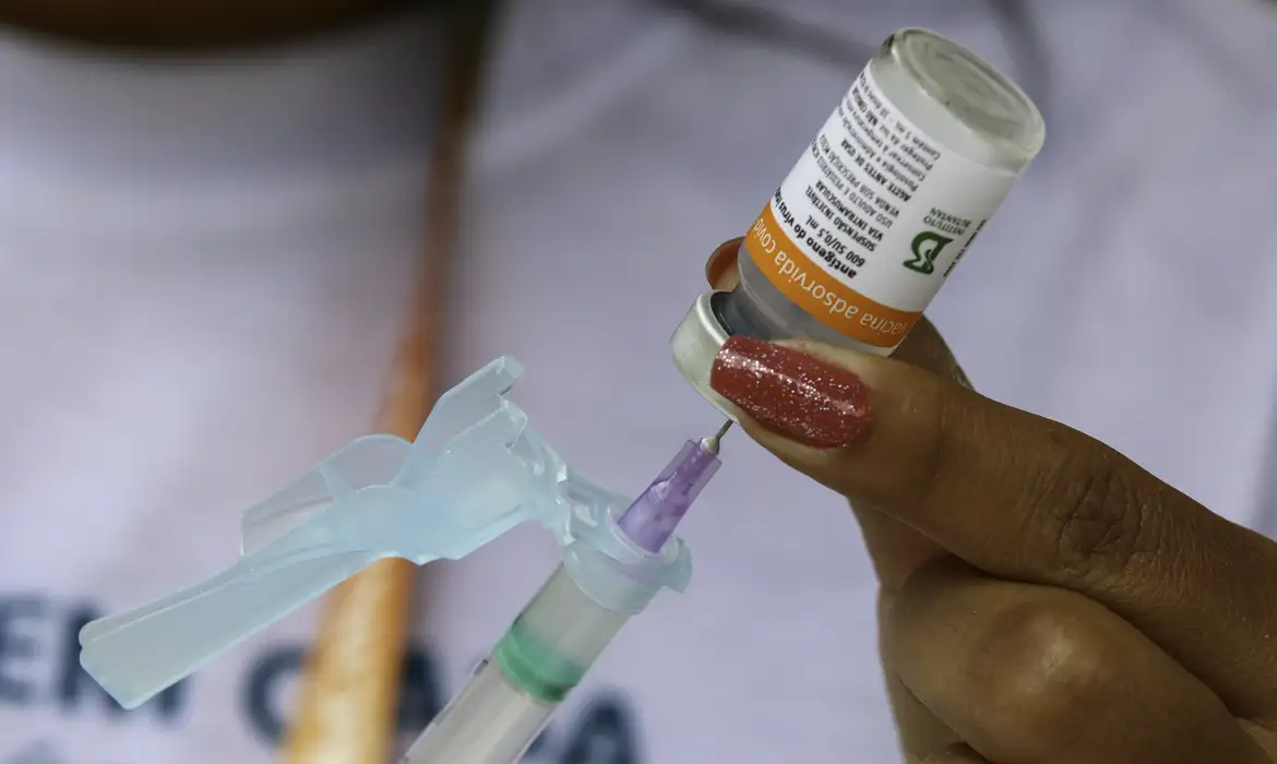 Mão feminina prepara seringa com dose de vacina CoronaVac, frasco em destaque