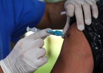 Profissional de saúde com luvas injetando vacina no braço de adulto de camiseta preta