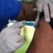 Profissional de saúde com luvas injetando vacina no braço de adulto de camiseta preta