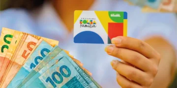 Beneficiário segura cartão do Bolsa Família ao lado de cédulas de dinheiro.