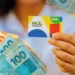 Beneficiário segura cartão do Bolsa Família ao lado de cédulas de dinheiro.
