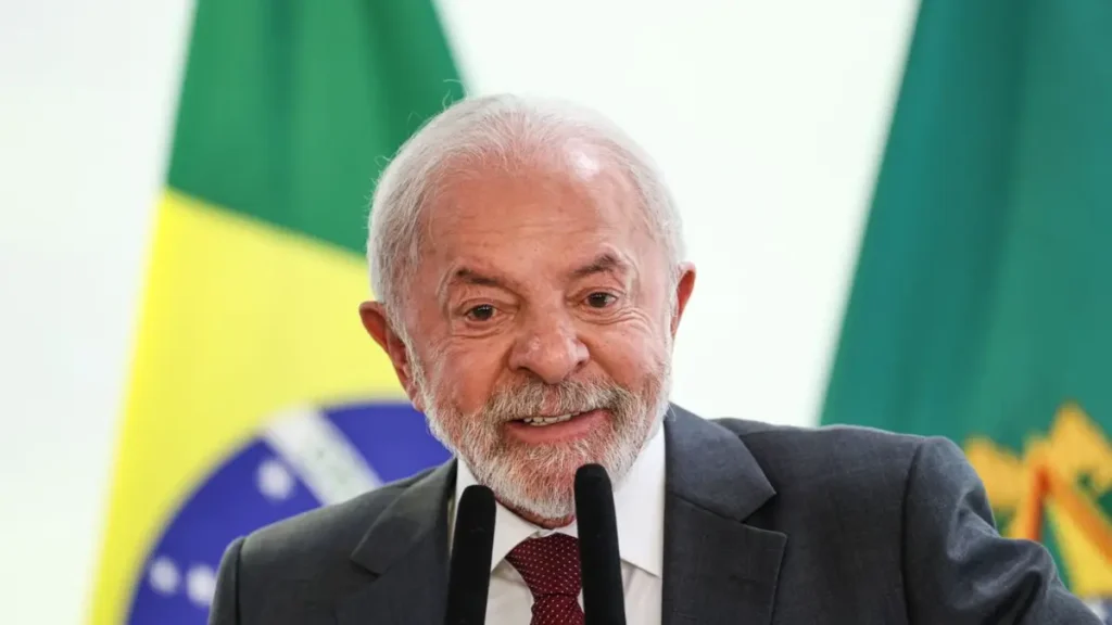 O presidente Luiz Inácio Lula da Silva discursa sorridente em frente a um microfone, com bandeiras ao fundo.