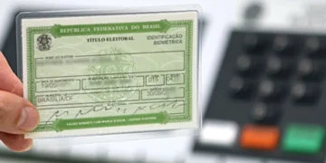 Mão segurando o Título de Eleitor com urna eletrônica ao fundo.