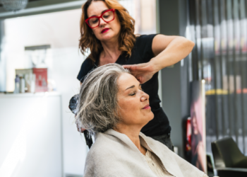 Mulher de cabelos grisalhos relaxa enquanto cabeleireira faz escova em salão moderno.
