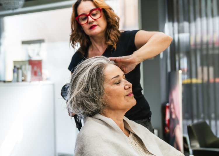 Mulher de cabelos grisalhos relaxa enquanto cabeleireira faz escova em salão moderno.