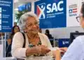 Mulher idosa sorridente entrega documento a uma atendente em balcão de atendimento prioritário, com sinalização do SAC ao fundo.