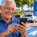 Idoso sorridente utiliza celular próximo a uma vaga de estacionamento reservada para pessoas com 60 anos ou mais.