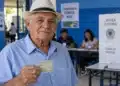 Idoso brasileiro com título de eleitor na mão, em uma seção eleitoral, com mesários e cabine de votação ao fundo.