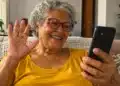 Idosa de cabelos grisalhos e óculos sorri enquanto participa de uma videochamada pelo celular, sentada no sofá de casa.