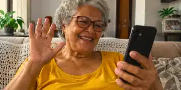 Idosa de cabelos grisalhos e óculos sorri enquanto participa de uma videochamada pelo celular, sentada no sofá de casa.