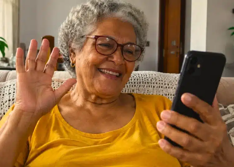 Idosa de cabelos grisalhos e óculos sorri enquanto participa de uma videochamada pelo celular, sentada no sofá de casa.