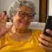 Idosa de cabelos grisalhos e óculos sorri enquanto participa de uma videochamada pelo celular, sentada no sofá de casa.