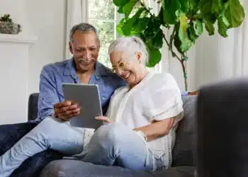 Casal de idosos sorrindo enquanto utiliza um tablet no sofá de casa.