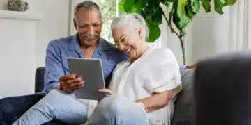 Casal de idosos sorrindo enquanto utiliza um tablet no sofá de casa.