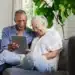 Casal de idosos sorrindo enquanto utiliza um tablet no sofá de casa.
