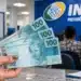 Mão segura notas de R$ 100 em agência do INSS, representando o pagamento da pensão por morte