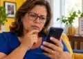 Mulher de meia-idade, usando óculos e blusa azul, observa o celular com expressão de dúvida, representando o acesso ao Meu INSS e ao INSS Digital