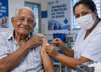 Idoso com mais de 85 anos recebe vacina no braço aplicada por profissional de enfermagem em unidade de saúde, ilustrando a vacinação contra pneumonia na Baixada Santista.