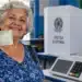 Idosa segura título de eleitor ao lado de urna eletrônica em seção eleitoral, representando o voto como comprovação da Prova de Vida do INSS.